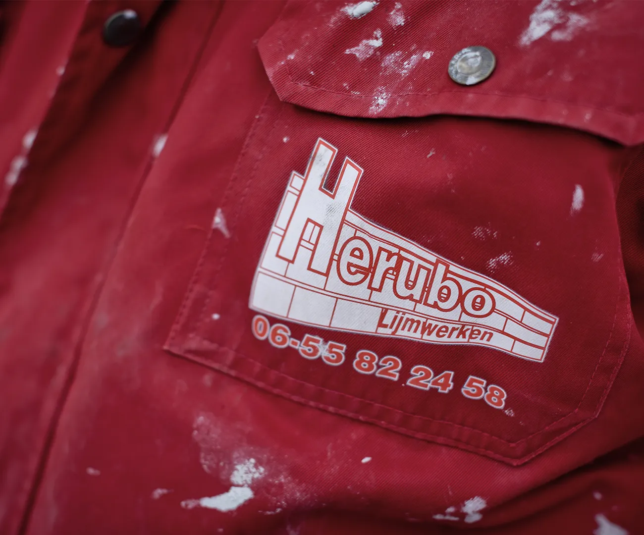 Close-up van het HERUBO-logo op de borstzak van een rode, met lijmspatten besmeurde werkjas