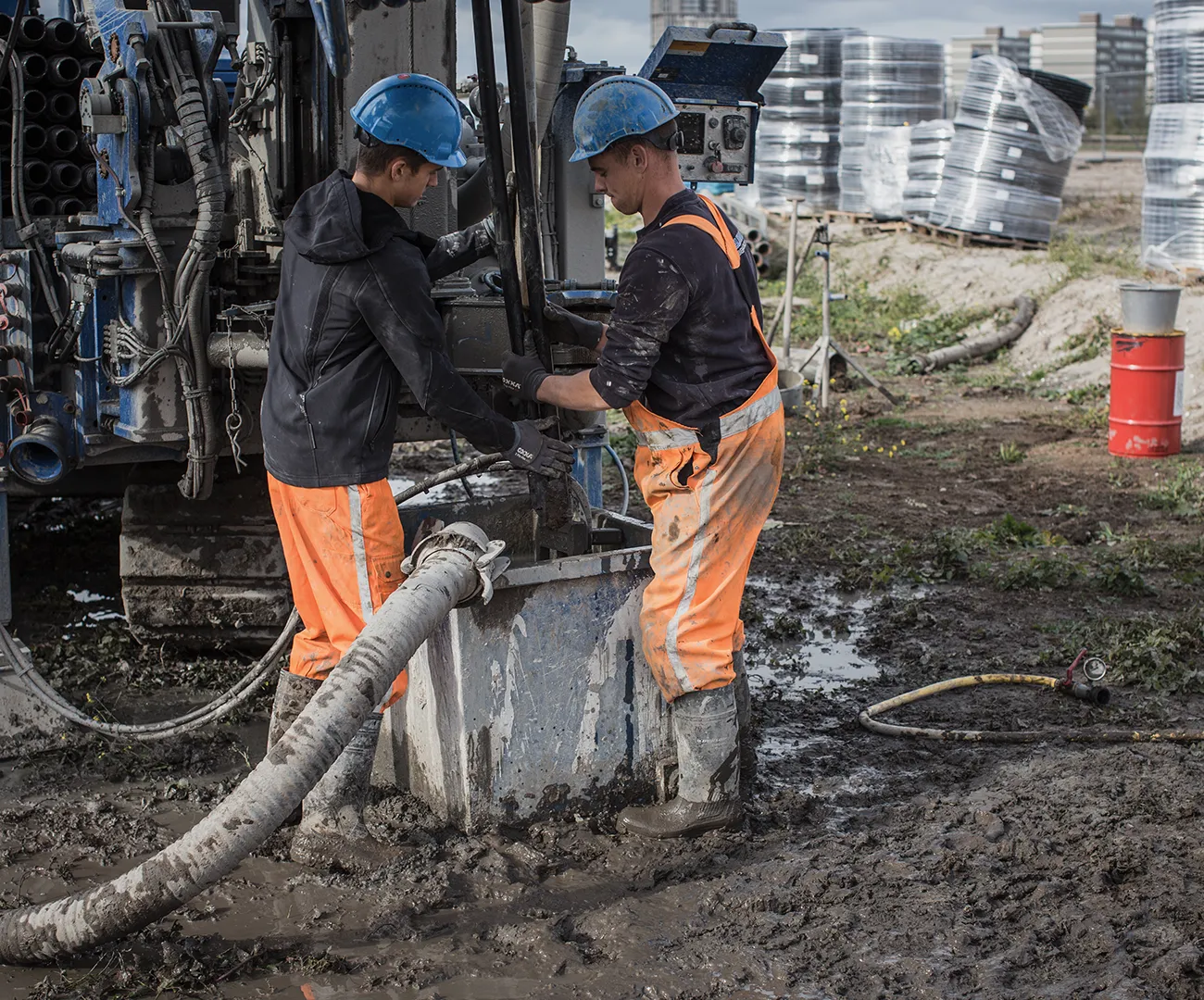 Twee werknemers van Koelewijn Bronbemalingen in oranje werkkleding en veiligheidshelm bedienen een boorinstallatie op een modderige bouwplaats
