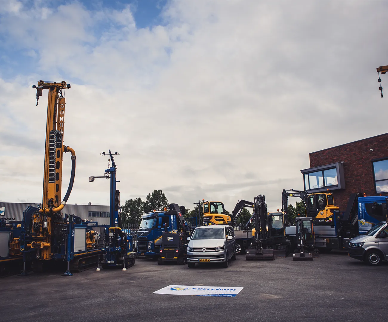 Materieel van Koelewijn Bronbemalingen opgesteld op het bedrijfsterrein: boorstellingen, graafmachines, vrachtwagens en bedrijfsbussen onder een bewolkte lucht