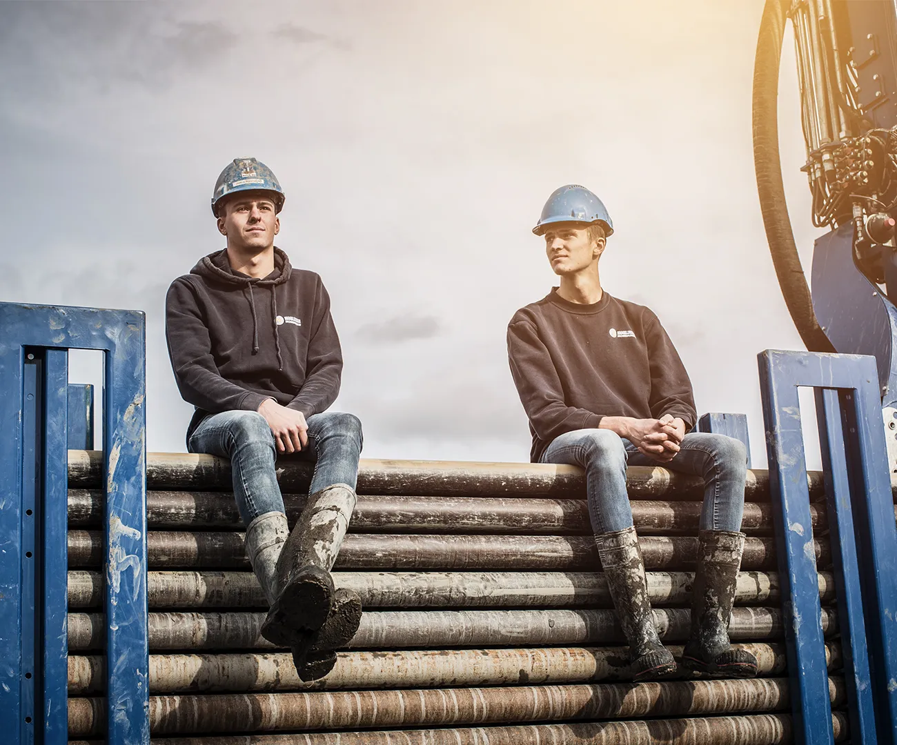 Twee werknemers van Koelewijn Bronbemalingen met veiligheidshelm en modderige laarzen, zittend op een stapel bemalingsbuizen bij een boorinstallatie