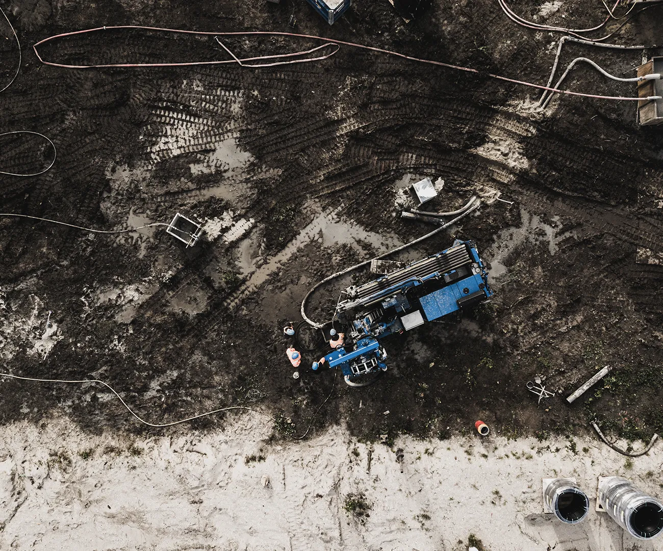 Luchtfoto van een bronbemalingsinstallatie op een modderige bouwplaats, met een blauwe boormachine, twee werknemers en aangesloten slangen en filters